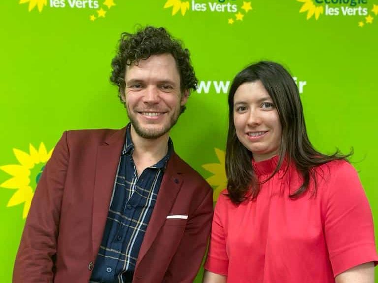 Morgan Jasienski et Alix Florent, deux jeunes personnes aux habits colorés, sourient devant un fond vert et jaune aux couleurs de EELV. Ce sont les deux responsable du pôle "projet et élus" du BED de EELV Paris